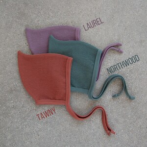 May include: Three waffle-knit baby bonnets in shades of tawny, Northwood, and laurel. Each bonnet has a matching trim and ties. The names of the colors are printed on the image.