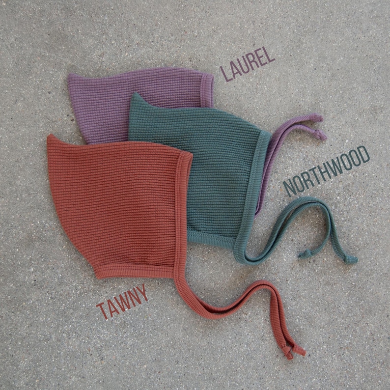 May include: Three waffle-knit baby bonnets in shades of tawny, Northwood, and laurel. Each bonnet has a matching trim and ties. The names of the colors are printed on the image.