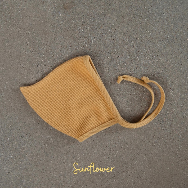 May include: A mustard yellow baby bonnet with a textured waffle knit design. The bonnet has a rounded shape with a brim and ties. The word "Sunflower" is written in a cursive font at the bottom of the image.