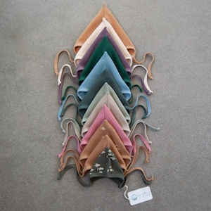 May include: A collection of colorful baby bonnets in various shades, including tan, purple, green, and blue. Each bonnet has a textured waffle weave and ties. The bonnets are arranged in a layered, symmetrical pattern.