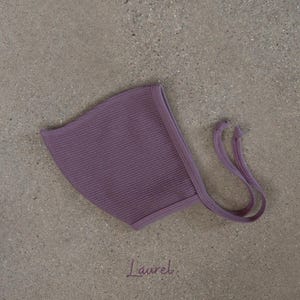 May include: A lavender-colored baby bonnet with a waffle knit texture. The bonnet has a rounded shape and features long, thin ties on each side. The word "Laurel" is written in cursive at the bottom of the image.