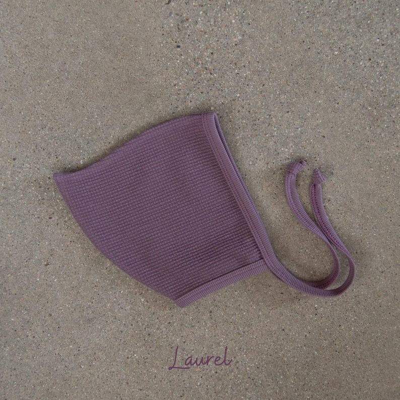 May include: A lavender-colored baby bonnet with a waffle knit texture. The bonnet has a rounded shape and features long, thin ties on each side. The word "Laurel" is written in cursive at the bottom of the image.