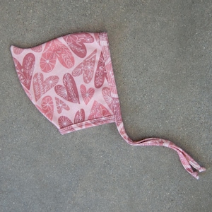 以下が含まれることがあります： 白いハートと花の模様がついたピンクのベビーボンネット