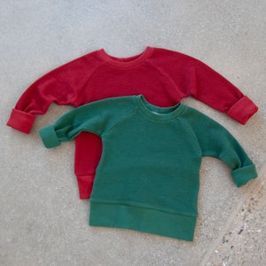 May include: Two long-sleeved waffle-knit shirts. One is a deep red, the other is a muted green. Both shirts have a crew neck and rolled cuffs. The shirts are laying on a gray concrete surface.