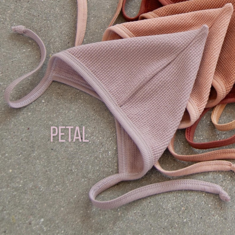 May include: A close-up of several baby bonnets in shades of pink and peach. The bonnet in the foreground is petal pink, with a textured waffle weave and ties. The word "PETAL" is printed in the lower left corner.