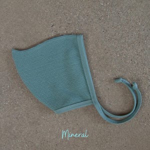May include: A light teal baby bonnet with a textured knit pattern. The bonnet has a rounded shape with a brim and ties. The word "Mineral" is written in a script font at the bottom of the image.