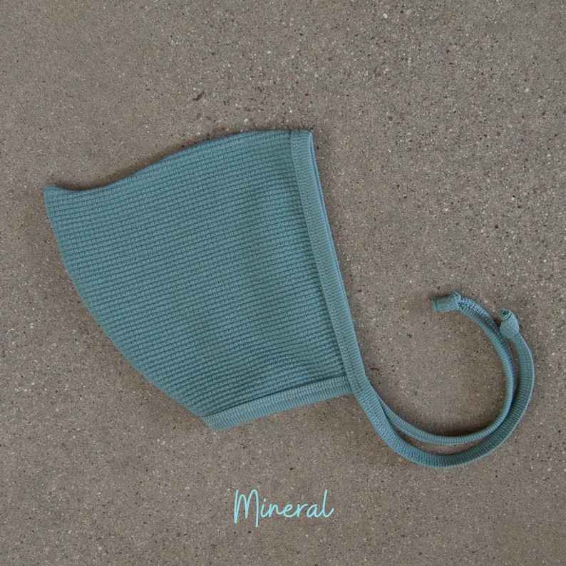 May include: A light teal baby bonnet with a textured knit pattern. The bonnet has a rounded shape with a brim and ties. The word "Mineral" is written in a script font at the bottom of the image.
