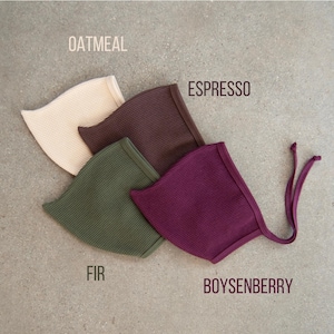 May include: Four waffle-knit baby bonnets in neutral colors: oatmeal, espresso, fir, and boysenberry. Each bonnet has a rounded shape and a tie closure. The names of the colors are printed above each bonnet.