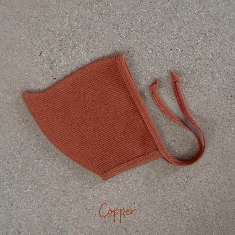 May include: A copper-colored baby bonnet with a waffle knit texture. The bonnet has a rounded shape with a brim and ties for securing. The word "Copper" is written in cursive at the bottom of the image.
