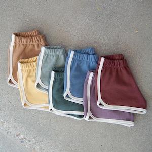 May include: A collection of six pairs of retro-style shorts in various colors, including brown, yellow, blue, green, purple, and maroon. Each pair features a white trim along the edges and a white stripe down the side.
