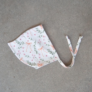 May include: A white baby bonnet with a floral pattern of pink bows, hearts, and green leaves. The bonnet has tie strings and is made of soft fabric. A charming accessory for a newborn.