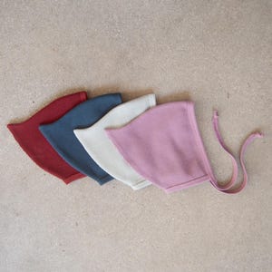 May include: Four ribbed knit baby bonnets in shades of red, blue, off-white, and pink. The bonnets are arranged in a row, with the pink bonnet featuring attached ties. The bonnets are made of a soft, textured fabric.