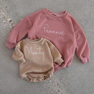 May include: A dusty rose long-sleeved sweatshirt with the name Frannie embroidered in white script and a tan long-sleeved romper with the name Monty embroidered in white script, featuring a round neckline and snap closures at the bottom.