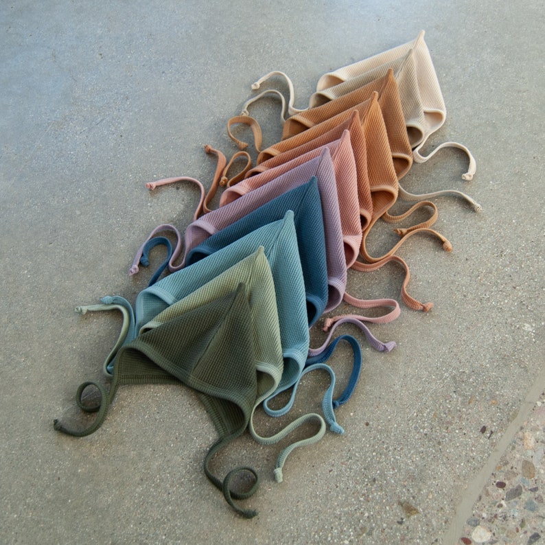May include: An assortment of baby bonnets in various colors, including olive green, teal, blue, lavender, pink, and tan. Each bonnet has a textured waffle weave and ties with matching fabric strings. The bonnets are arranged in a diagonal line.