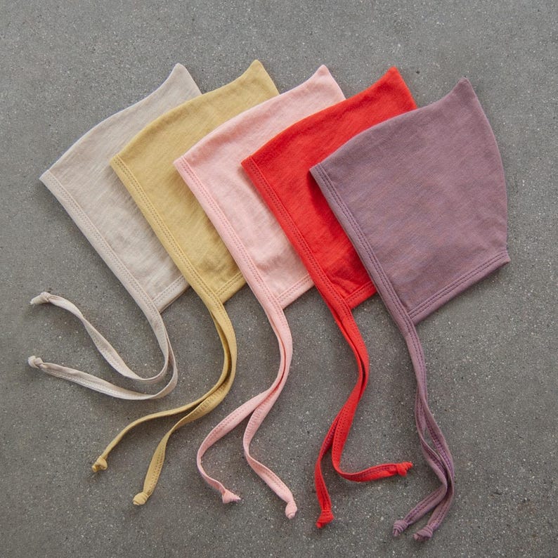 May include: Five pastel-colored baby bonnets arranged diagonally. The bonnets are in shades of beige, yellow, pink, red, and purple. Each bonnet has long, thin ties with small knots at the ends. The bonnets are made of a soft, lightweight fabric.