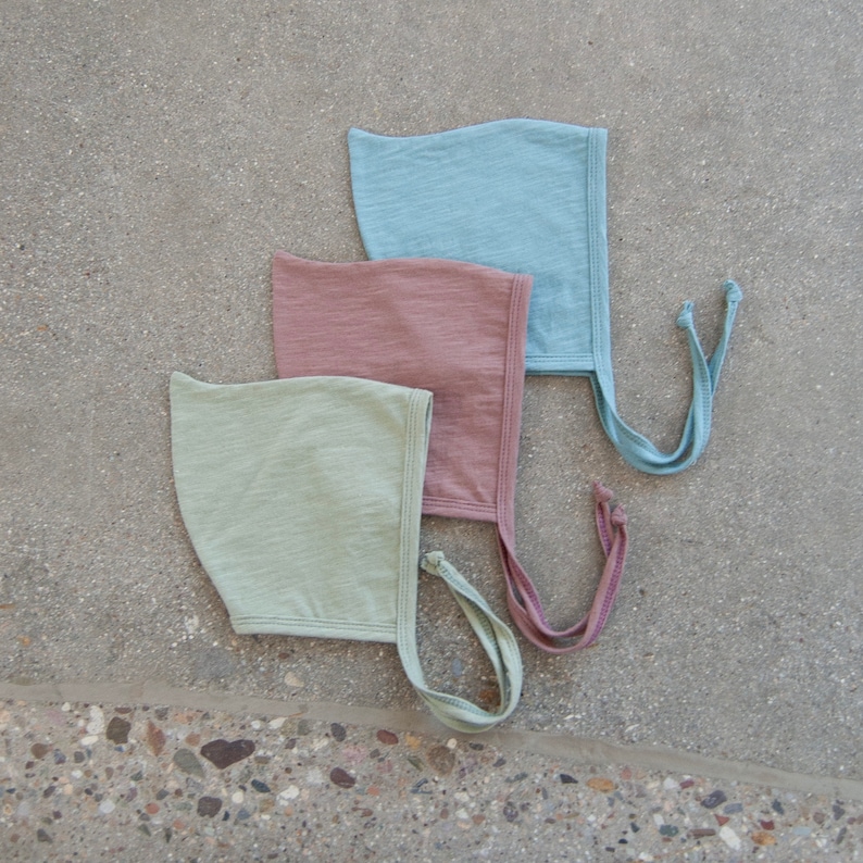 May include: Three baby bonnets in pastel colors: light green, dusty rose, and light blue. Each bonnet has a rounded shape with a tie closure. The bonnets are made of a soft, textured fabric.