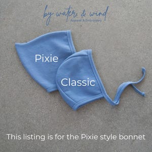 May include: Two light blue baby bonnets, one labeled "Pixie" and the other "Classic." The bonnets are made of soft fabric and have a simple design. The text "by water & wind Apparel & Embroidery" is at the top. The text "This listing is for the Pixie style bonnet" is at the bottom.