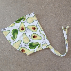 May include: White baby bonnet with a repeating pattern of green and yellow avocados, some halved to show the brown pit. The bonnet has a tie closure and is made of soft fabric.