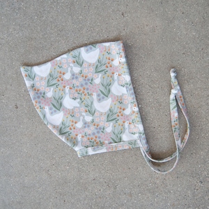 Puede incluir: Un gorro de bebé con un estampado floral y de gansos. El gorro es de color gris claro con un patrón repetido de gansos blancos, pequeñas flores y hojas verdes. Tiene lazos a cada lado.