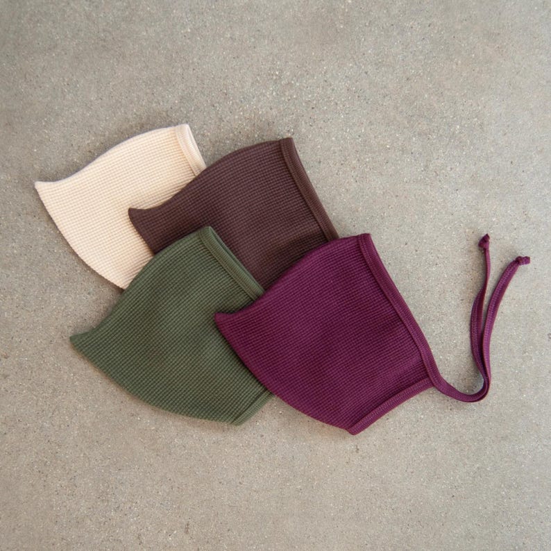 May include: Four waffle-knit baby bonnets in neutral colors: cream, brown, olive green, and deep purple. The bonnets have a rounded shape with a brim and ties. The bonnets are arranged in a diagonal pattern on a gray surface.