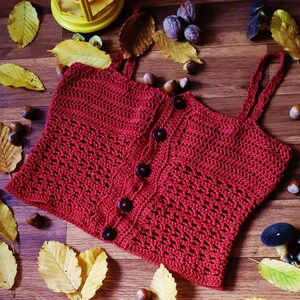 Puede incluir: Un top de crochet rojo con cierre de bot&oacute;n y tirantes finos. El top est&aacute; colocado sobre una superficie de madera con hojas de oto&ntilde;o y bellotas esparcidas a su alrededor.