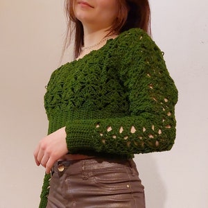Op de afbeelding: Een groene gehaakte trui met een kantpatroon. De trui heeft lange mouwen en een licht ingekorte lengte.