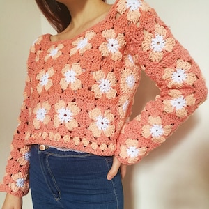 May include: A crocheted sweater with a floral pattern in shades of peach and white. The sweater has a loose fit and long sleeves.