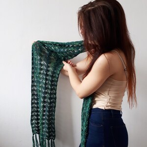 May include: A green and blue crocheted scarf with fringe tassels. The scarf is draped over the shoulder of a person wearing a beige tank top and blue jeans.