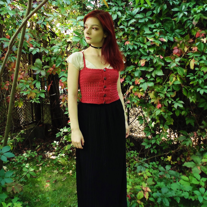 Puede incluir: Una mujer con un top corto de crochet rojo con botones y una falda maxi negra. Est&aacute; de pie frente a un fondo verde frondoso.