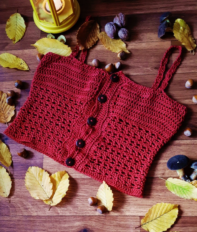 Puede incluir: Un top de crochet rojo con cierre de bot&oacute;n y tirantes finos. El top est&aacute; colocado sobre una superficie de madera con hojas de oto&ntilde;o y bellotas esparcidas a su alrededor.
