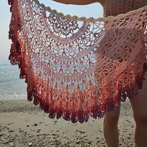 May include: A handmade, open-weave shawl in shades of pink and red, with a scalloped edge. The crochet shawl is held up against a backdrop of the ocean and sky, showcasing its intricate floral design and gradient colour.