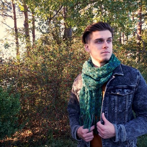 May include: A man wearing a denim jacket with a green and blue crocheted scarf. He is standing in front of a green bush.