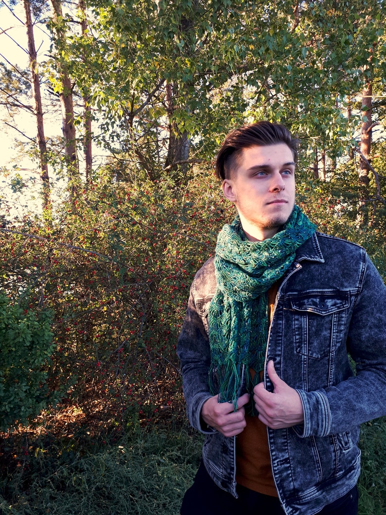 May include: A man wearing a denim jacket with a green and blue crocheted scarf. He is standing in front of a green bush.