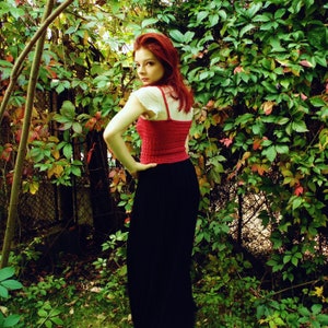Puede incluir: Una mujer con el pelo rojo lleva un top de crochet rojo y una falda negra. Est&aacute; de pie frente a una pared de enredaderas verdes.