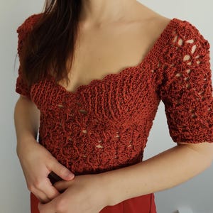 Puede incluir: Un top de ganchillo con cuello en V profundo y mangas cortas. El top está hecho de hilo de color óxido y tiene un delicado patrón de encaje.