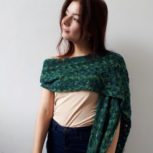 May include: A woman wearing a green and blue crocheted shawl with a beige tank top and blue jeans.