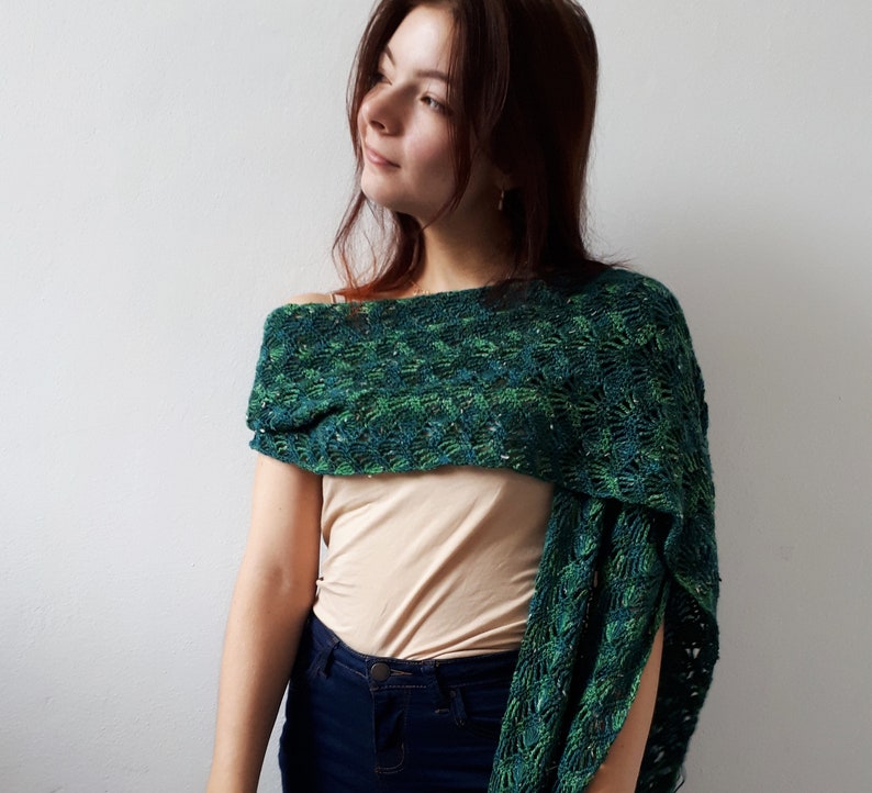 May include: A woman wearing a green and blue crocheted shawl with a beige tank top and blue jeans.