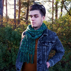 May include: A man wearing a denim jacket with a green and blue scarf. The scarf has a fringed edge. The man is standing in a forest setting.