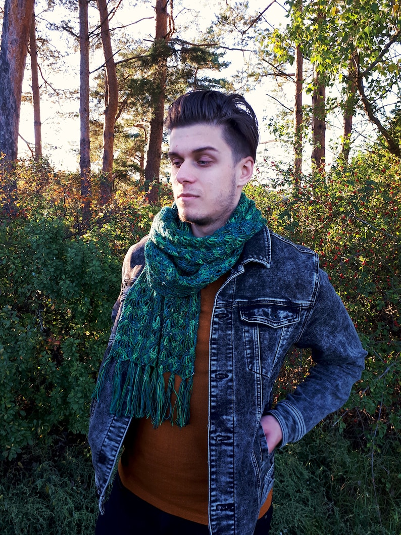 May include: A man wearing a denim jacket with a green and blue scarf. The scarf has a fringed edge. The man is standing in a forest setting.