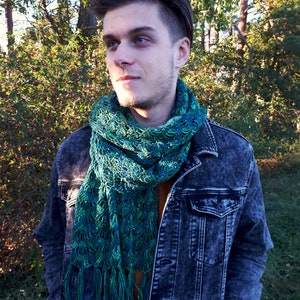 May include: A man wearing a denim jacket with a green and blue crocheted scarf with fringe.
