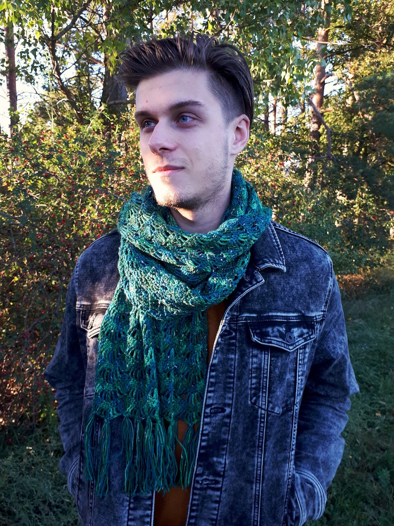May include: A man wearing a denim jacket with a green and blue crocheted scarf with fringe.
