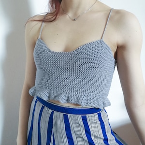 Puede incluir: Un top corto de crochet gris con un dobladillo con volantes. El top tiene tirantes finos y un escote de corazón.