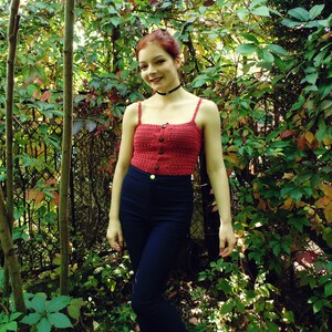 Puede incluir: Una mujer con un top de crochet rojo con cierre de bot&oacute;n y un collar negro. Est&aacute; de pie frente a un fondo verde con hojas.