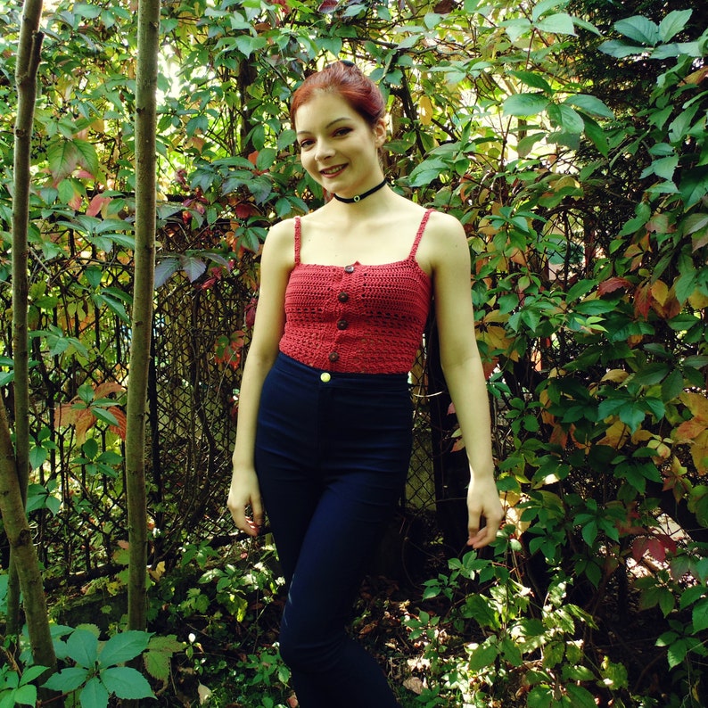 Puede incluir: Una mujer con un top de crochet rojo con cierre de bot&oacute;n y un collar negro. Est&aacute; de pie frente a un fondo verde con hojas.