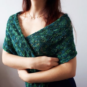 May include: A green and blue crocheted shawl with a delicate floral pattern. The shawl is draped over a person's shoulders and is held in place by their hands.