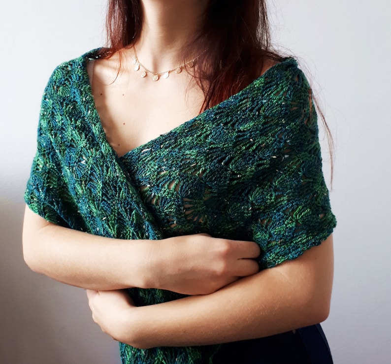 May include: A green and blue crocheted shawl with a delicate floral pattern. The shawl is draped over a person's shoulders and is held in place by their hands.