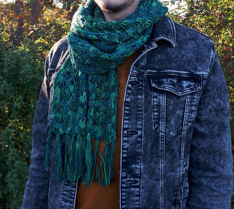 May include: A green and blue crocheted scarf with fringe. The scarf is worn around the neck of a person wearing a denim jacket.
