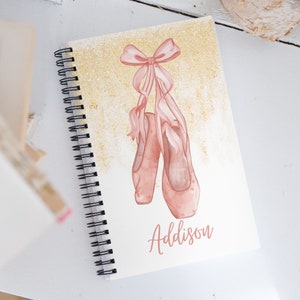 May include: A pink spiral notebook with a gold glitter background. The notebook features a watercolour illustration of pink ballet shoes with a pink ribbon. The name Addison is written in pink cursive on the front of the notebook.