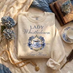 May include: Cream-colored sweatshirt with the text "LADY Whistledown Tea House" in blue script. The design includes a teacup illustration within a blue floral wreath. The sweatshirt is surrounded by a cozy aesthetic with books, flowers, and a teacup.