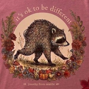 Könnte beinhalten: Ein malvenfarbenes T-Shirt mit einer Grafik eines Waschbären, der durch ein Feld läuft, umgeben von einem kreisförmigen Kranz aus Herbstblumen, Blättern und Kürbissen. Der Text lautet "it's ok to be different" und "jimothy from seattle".